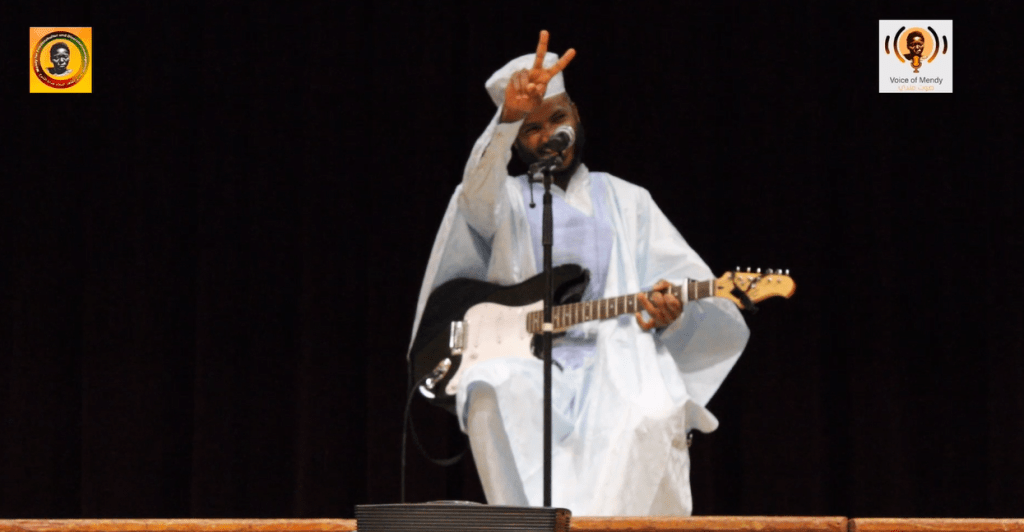 Ebo Krdum, the Sudanese-Swedish musician, is sitting with his guitar and holding a peace sign with his left hand.